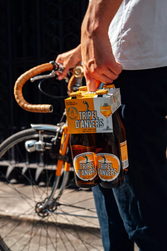 Foto van een man, met in de ene hand een pakket Tripel D'Anvers bier en de andere hand een oude koersfiets.